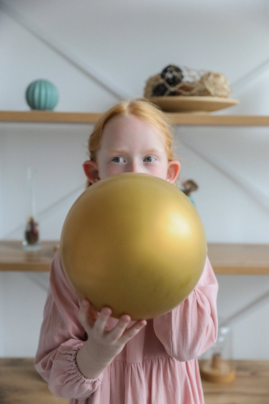 adorable child in girlish dress inflating balloon at home